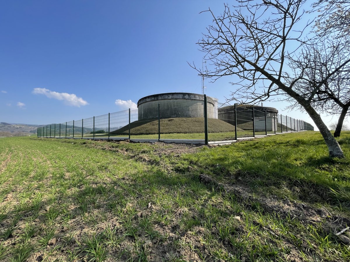 Cloture rigide a panneaux soudes verts ceinturant un site de reservoirs d'eau, sur fond de collines et ciel bleu