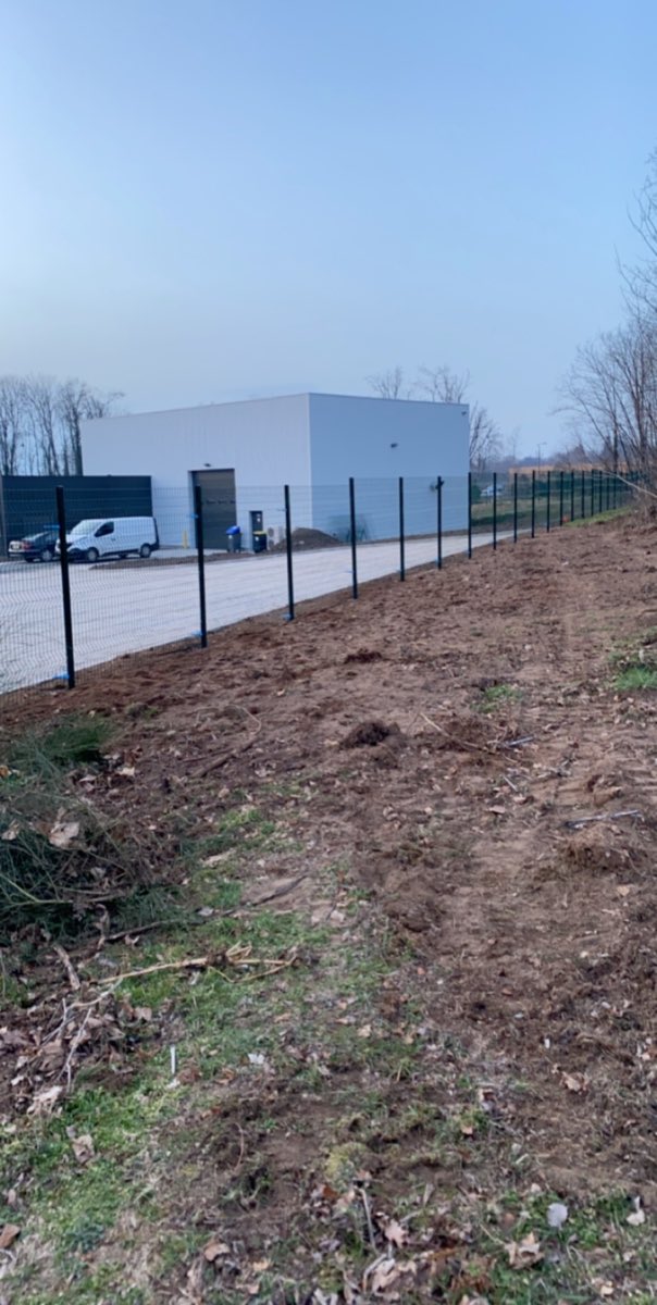 Cloture rigide a panneaux soudes anthracite sur poteaux, delimitant un site industriel avec batiment et vehicule utilitaire en arriere-plan