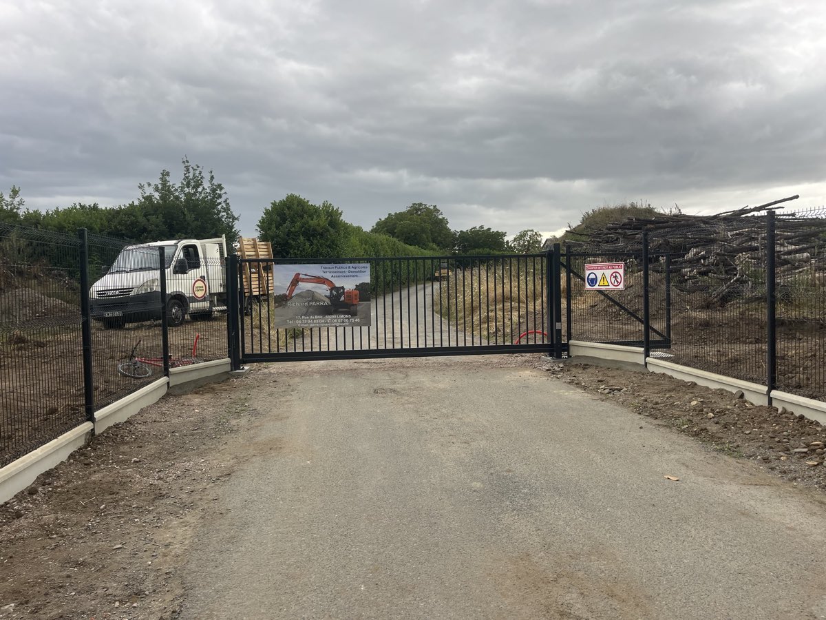 Portail autoportant coulissant noir a barreaudage vertical en position fermee, avec panneau de signalisation de chantier, raccorde a une cloture rigide sur longrines beton